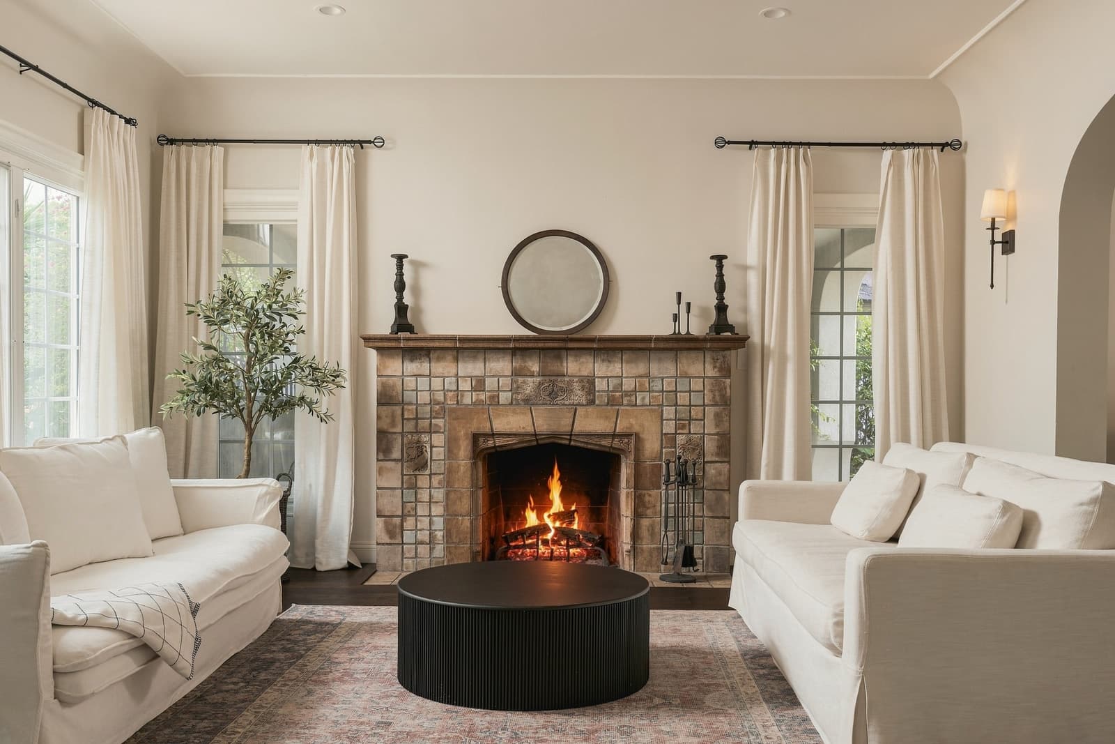 Living room with a tile fireplace lit, framed by cream sofas and full-length curtains